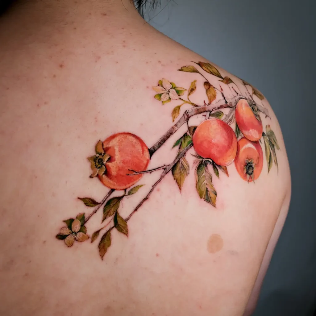 Colorful watercolor tattoo of a pomegranate branch with fruit and leaves, inked on a woman’s upper shoulder.