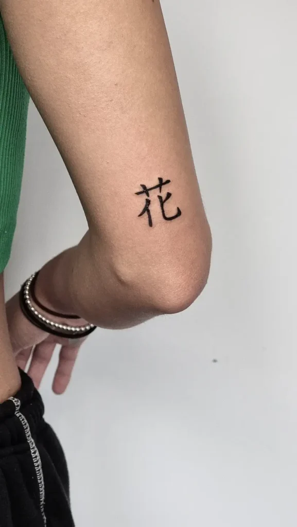 Black kanji character tattoo meaning “flower” inked above a woman’s elbow.