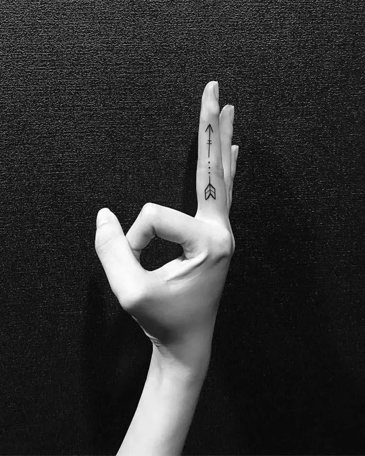 Black and white photo showing a woman’s hand in an "OK" gesture with a sleek arrow tattoo running along the middle finger. The minimalist tattoo features clean black lines and geometric elements, adding a modern and symbolic touch. Set against a textured dark background, the hand’s elegant pose and sharp contrast emphasize the tattoo’s bold yet refined style, perfect for finger tattoo inspiration.









