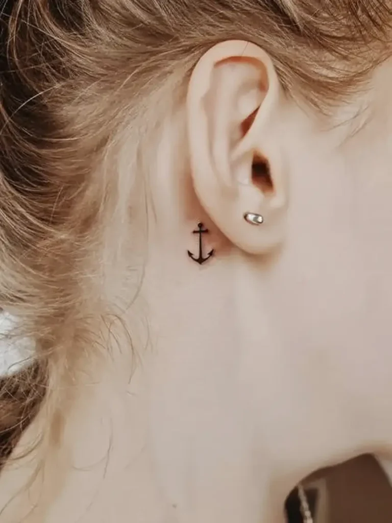 A minimalist black anchor tattoo placed behind a woman’s ear, featuring clean lines and simple design, photographed in soft lighting to highlight the subtle and symbolic placement.









