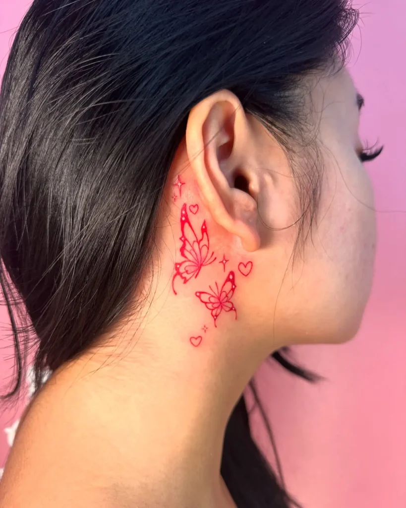 A behind the ear tattoo in bright red ink featuring two butterflies, small hearts, and sparkles, placed on a woman’s neck with long dark hair and a soft pink background.









