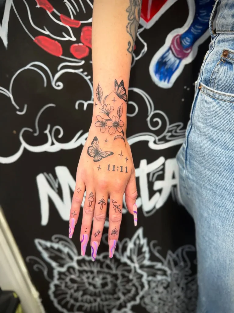 A hand with a butterfly and floral tattoo, featuring "11:11," stars, and finger symbols, paired with pink nails against a colorful background.
