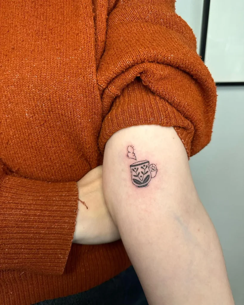 Small tattoo of a decorated coffee cup with steam shaped like a heart, inked on a woman’s upper arm.