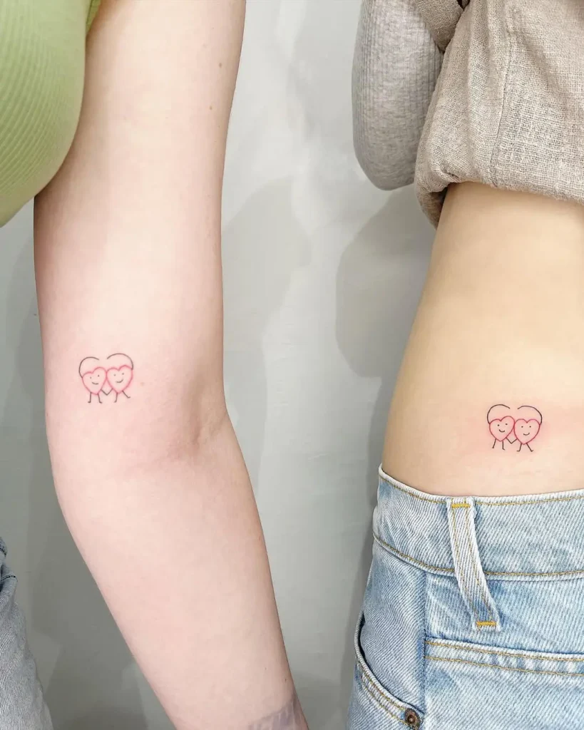 Matching pink heart tattoos with smiling faces and legs, symbolizing friendship and happiness in a minimal fine line style.