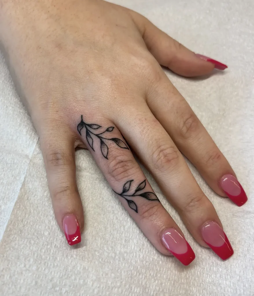 Close-up of a woman's hand with a fine-line leaf tattoo wrapping around the middle finger. The black ink botanical design is delicate and feminine, adding a natural and elegant touch to the hand. Her nails are styled with glossy red French tips on a pink base, creating a bold contrast against the soft tattoo. This finger tattoo idea is perfect for women who love nature-inspired ink with a modern nail art twist.








