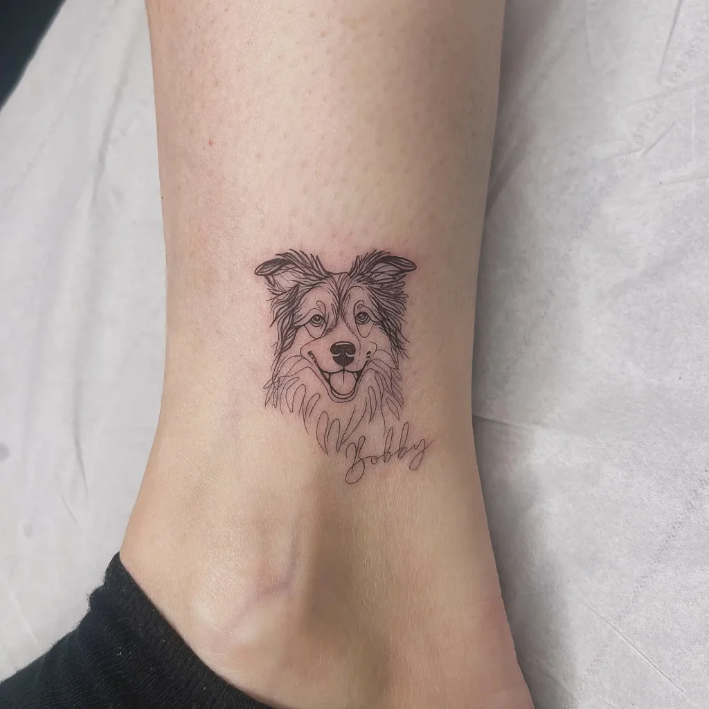 Minimal black line dog portrait tattoo near the ankle, showing a happy dog face with the name “Bobby” written below in cursive. Light, elegant ink style ideal for animal tattoo ideas simple and personal. A loving tribute for pet owners wanting emotional and customized ink in a small, visible placement.







Collage of four realistic dog portrait tattoos: one fluffy white dog, a pit bull on a hand, a small terrier with a toy, and a smiling dog framed by floral blooms. Done in black and grey realism style. Emotional, detailed pet tribute tattoos. Ideal for animal tattoo ideas simple yet personalized, showcasing loyalty and love. Great for arm, hand, or leg placements.









