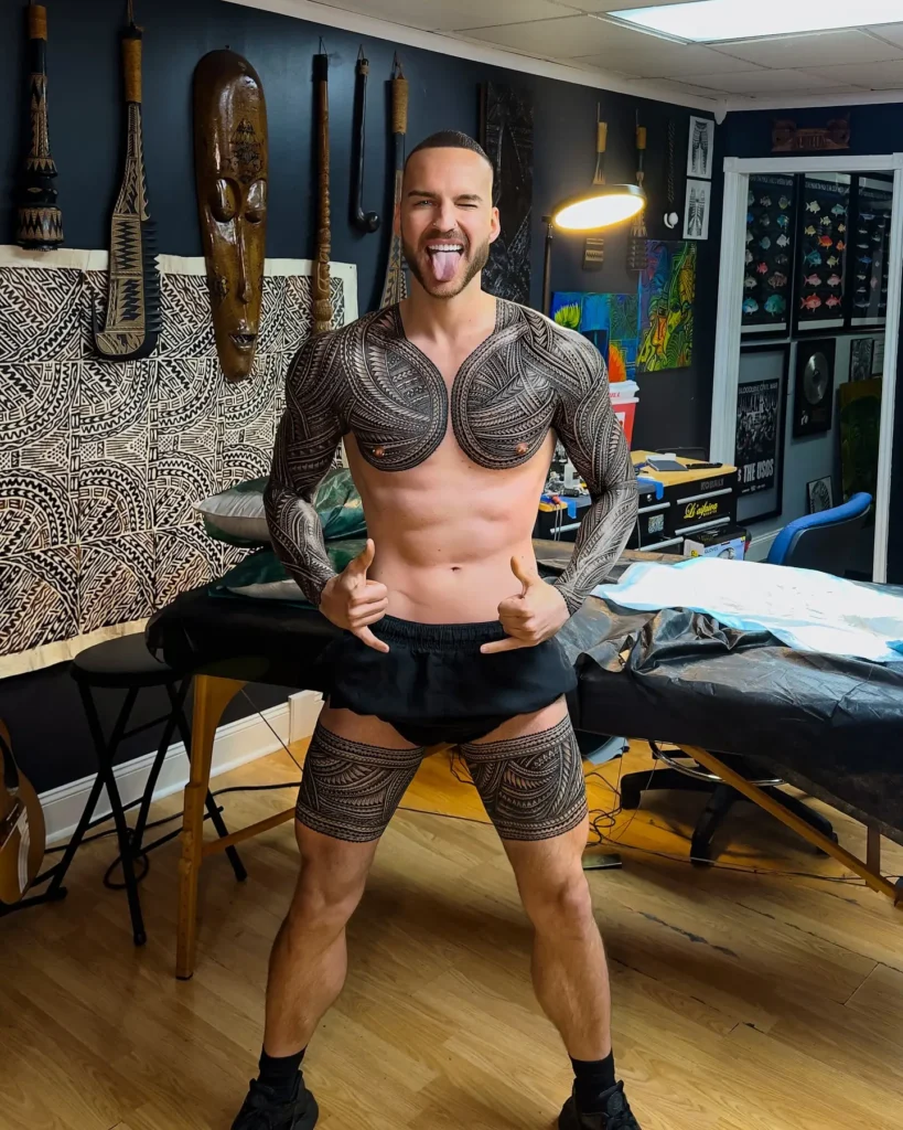 Polynesian-style chest tattoo for men featuring two large symmetrical shield-like patterns on the pectorals with intricate tribal detailing extending into full arm sleeves; traditional black ink design representing strength and cultural heritage.