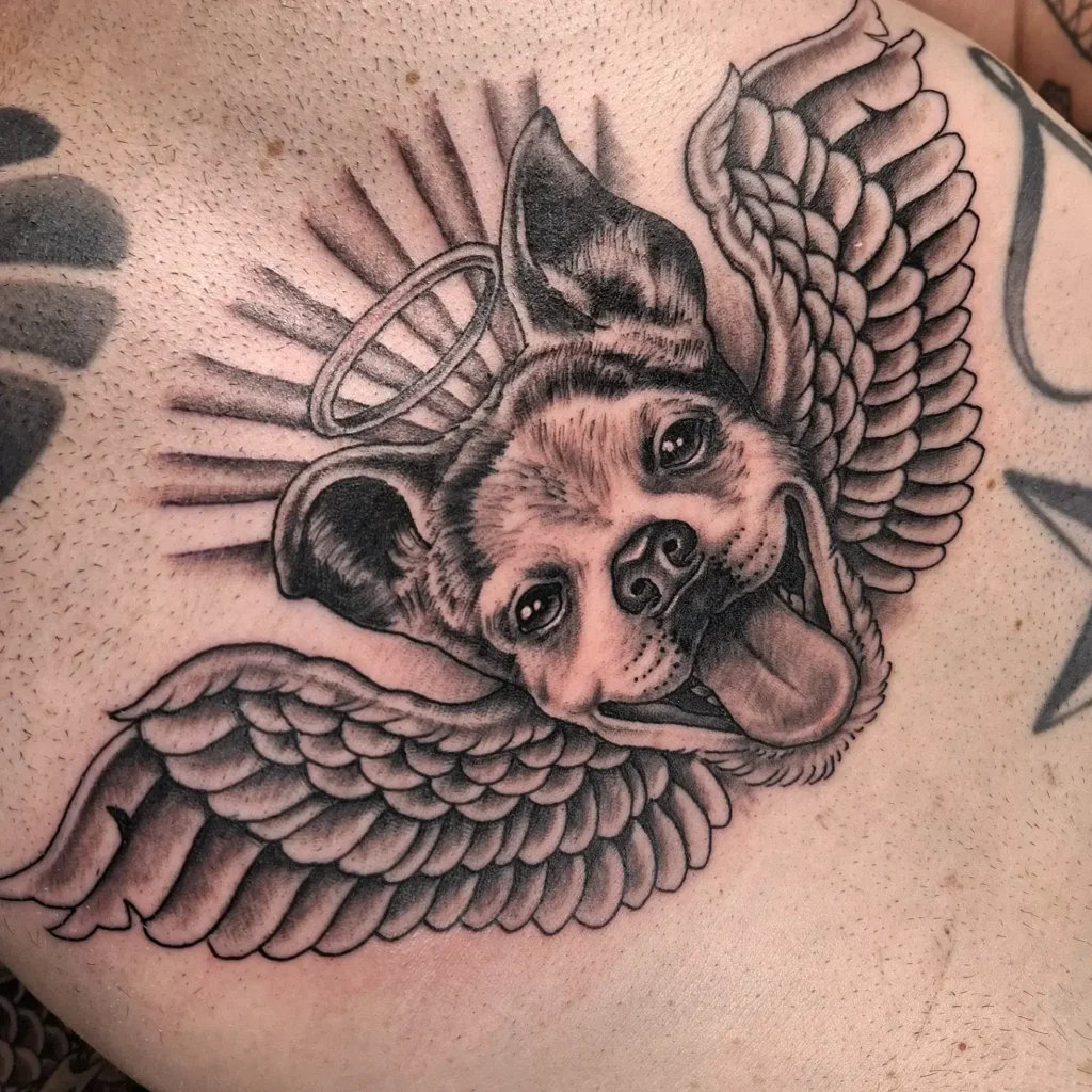  Black and gray tattoo of a smiling dog with angel wings and a halo, featuring radiant rays, designed as a heartfelt dog tattoo ideas memorial.