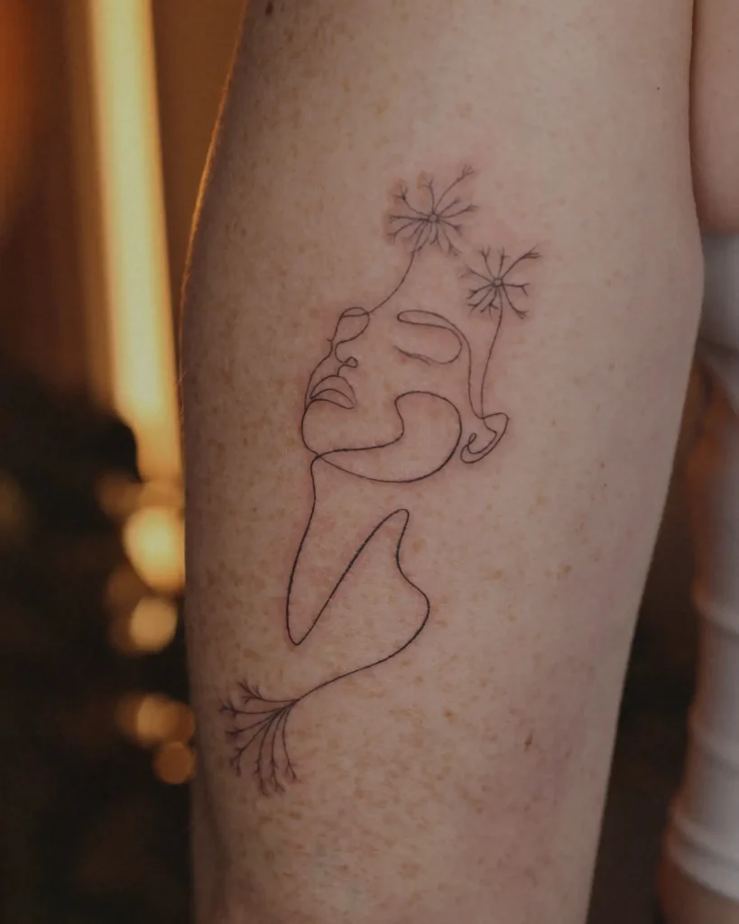 Minimalist line art tattoo of a woman’s face with dandelion flowers growing from her head, symbolizing peace and growth, placed on the thigh.

