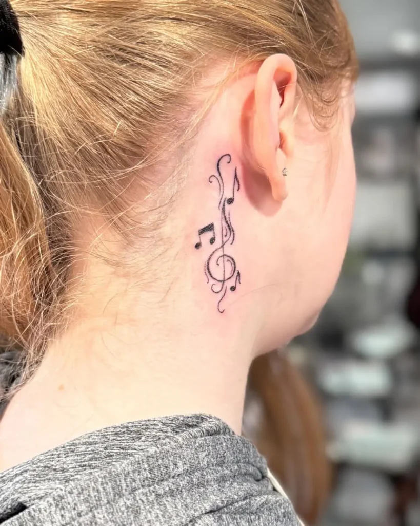 A behind the ear tattoo featuring black musical notes and a treble clef with swirling decorative lines, inked on a woman with light red hair tied back, highlighting the rhythm-inspired design.









