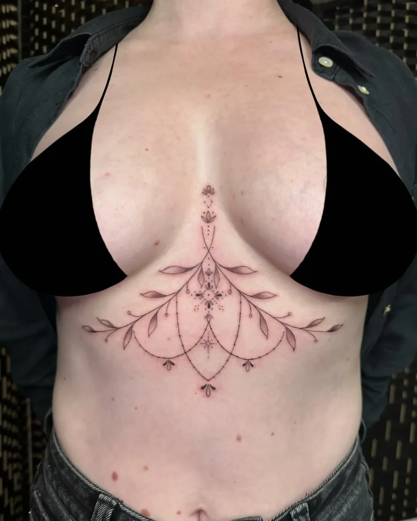 Ornamental underbust tattoo with floral motifs, leaves, and dotwork, inked in fine lines on a woman’s chest.