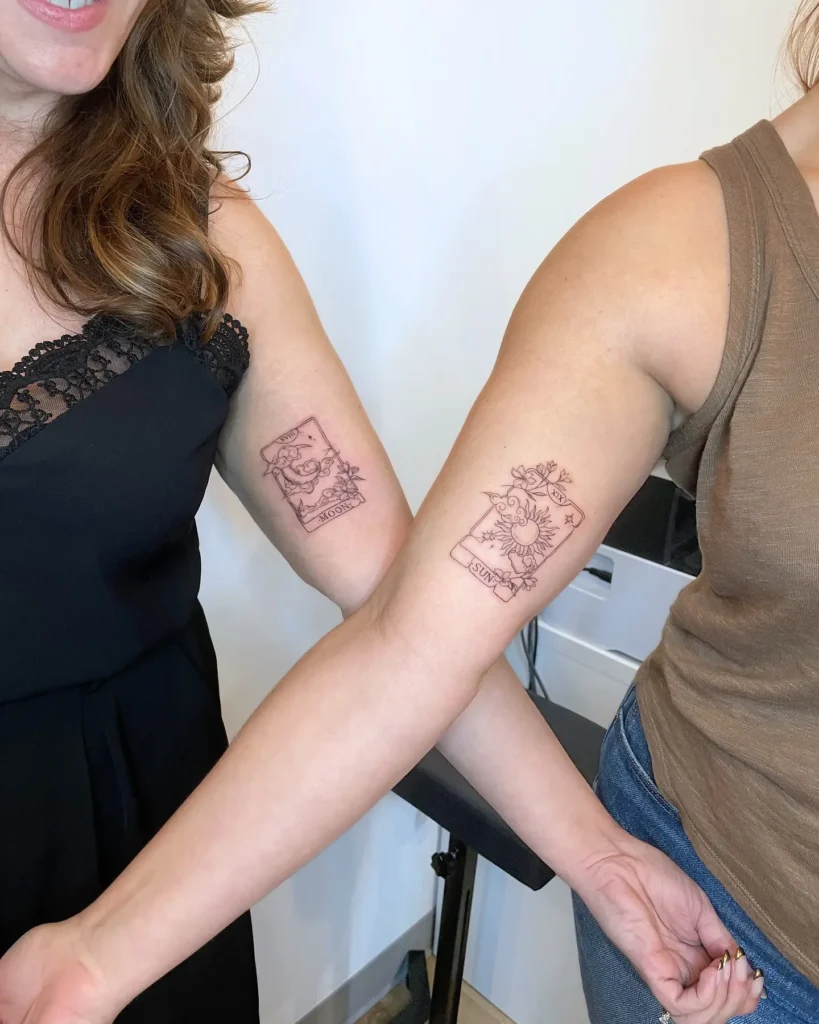 Matching arm tattoos show tarot card designs, one with a crescent moon and floral details labeled “MOON,” and the other with a radiant sun and flowers labeled “SUN.” The delicate linework adds elegance. These tattoos highlight mom and daughter tattoos unique, using sun and moon imagery to represent balance, connection, and the beauty of their relationship.